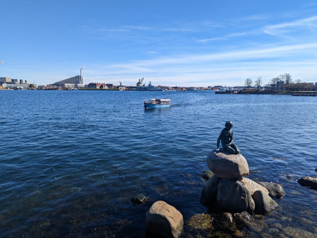 Statue of the Little Mermaid in front of the water and a boat in Copenhagen
